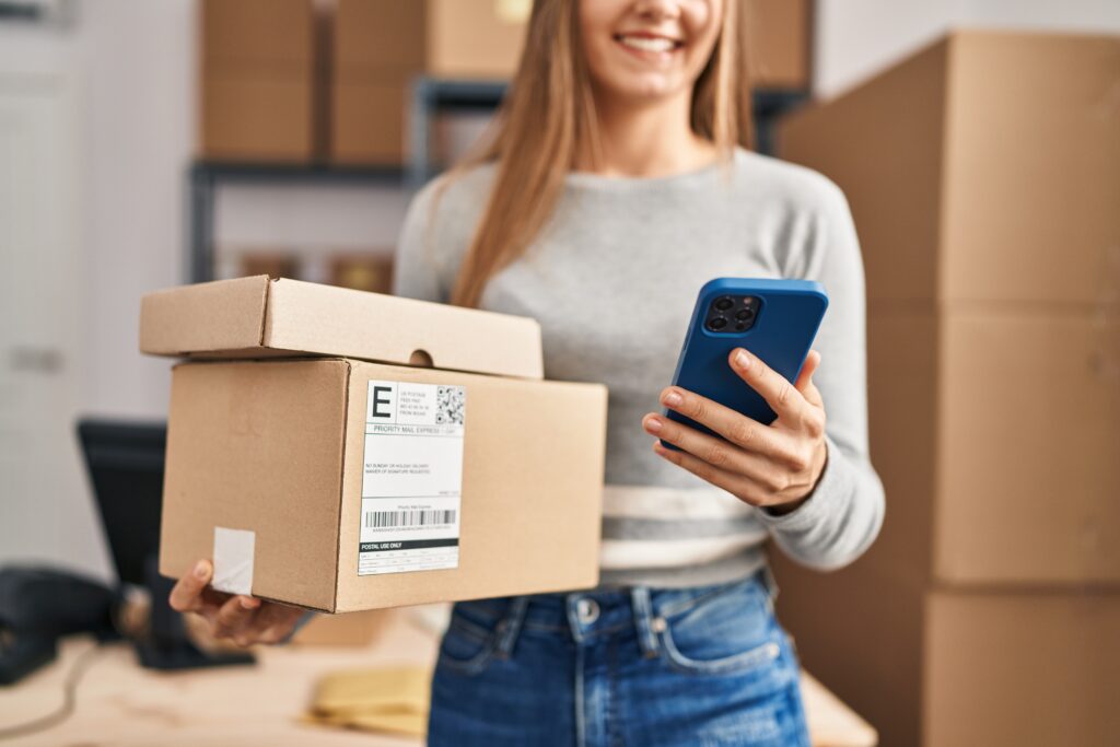 Young blonde woman ecommerce business worker using smartphone holding packages at office